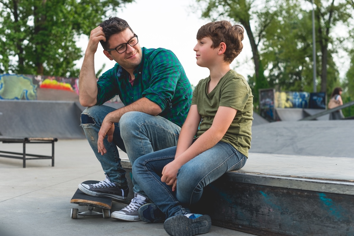 Father and pre-teen son sitting together at a skatepark